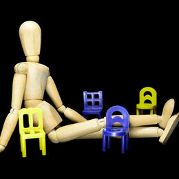 Holzpuppe auf dem Boden sitzend zwischen 6 kleinen bunten Stühlen