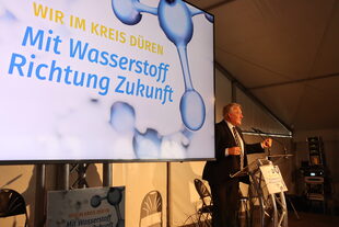 Landrat Wolfgang Spelthahn auf einer Bühne vor einem Bildschirm mit der Aufschrift "Mit Wasserstoff Richtung Zukunft".