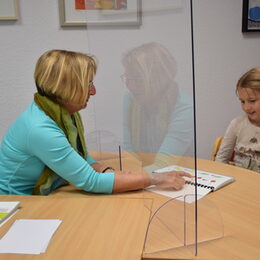 Birgit Bauer sitzt gegenüber von einem Kind am Schreibtisch und zeigt auf ein Buch mit Bildern.