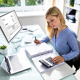 Junge Business-Frau sitzt am Schreibtisch. Vor ihr befinden sich ein Laptop, ein Ordner, ein Taschenrechner und ein Bildschirm.