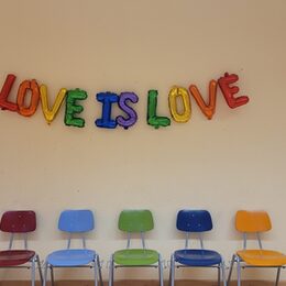 Buchstabenballons mit dem Schriftzug Love is Love an einer Wand über einer bunten Stuhlreihe