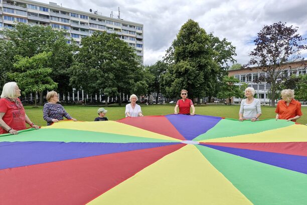Senioren und Kinder stehen zusammen im Kreis und schwingen ein großes Tuch in bunten Farben.