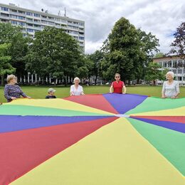 Senioren und Kinder stehen zusammen im Kreis und schwingen ein großes Tuch in bunten Farben.